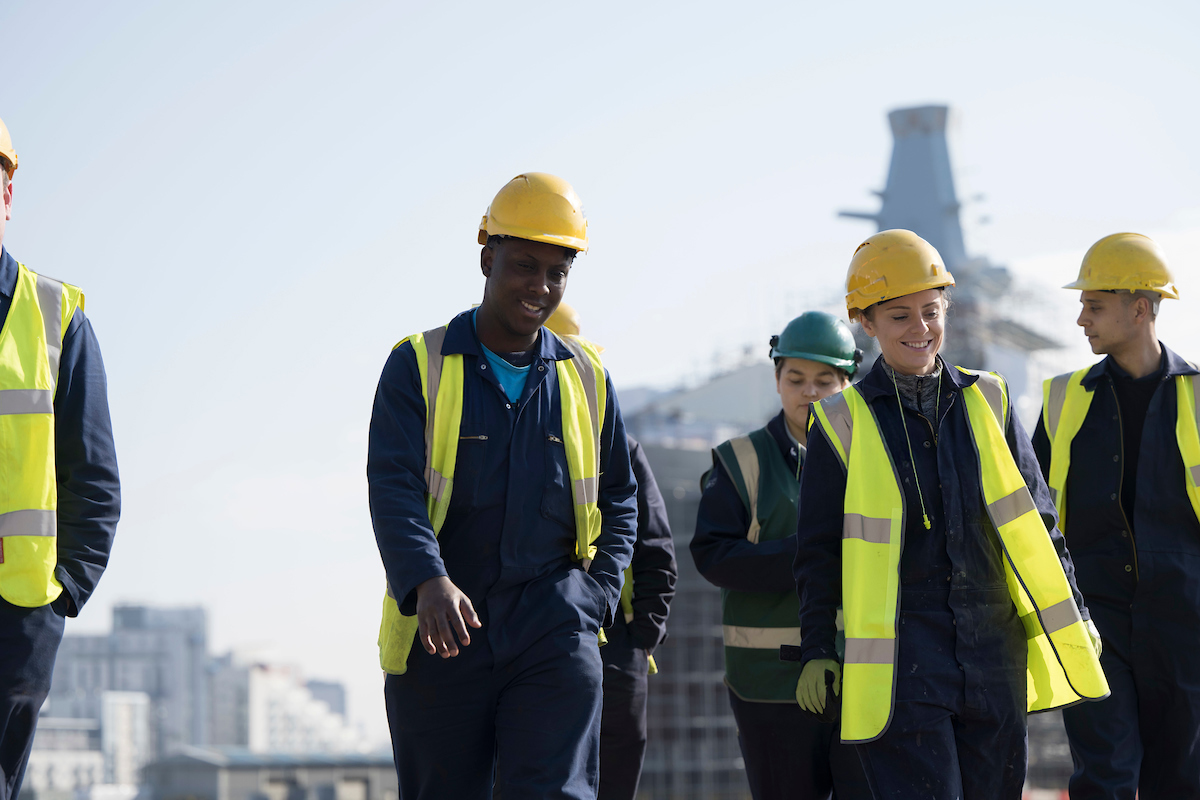 Apprentices in high-vis vests and hardhats walking with ship in the background